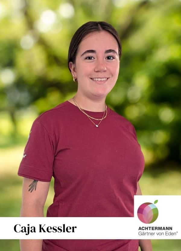 Caja Kessler, Porträt im Garten mit Achtermann-Gärtner-von-Eden-Logo Simon Huber trägt ein rotes T-Shirt mit dem Logo von Achtermann und steht mit verschränkten Armen.